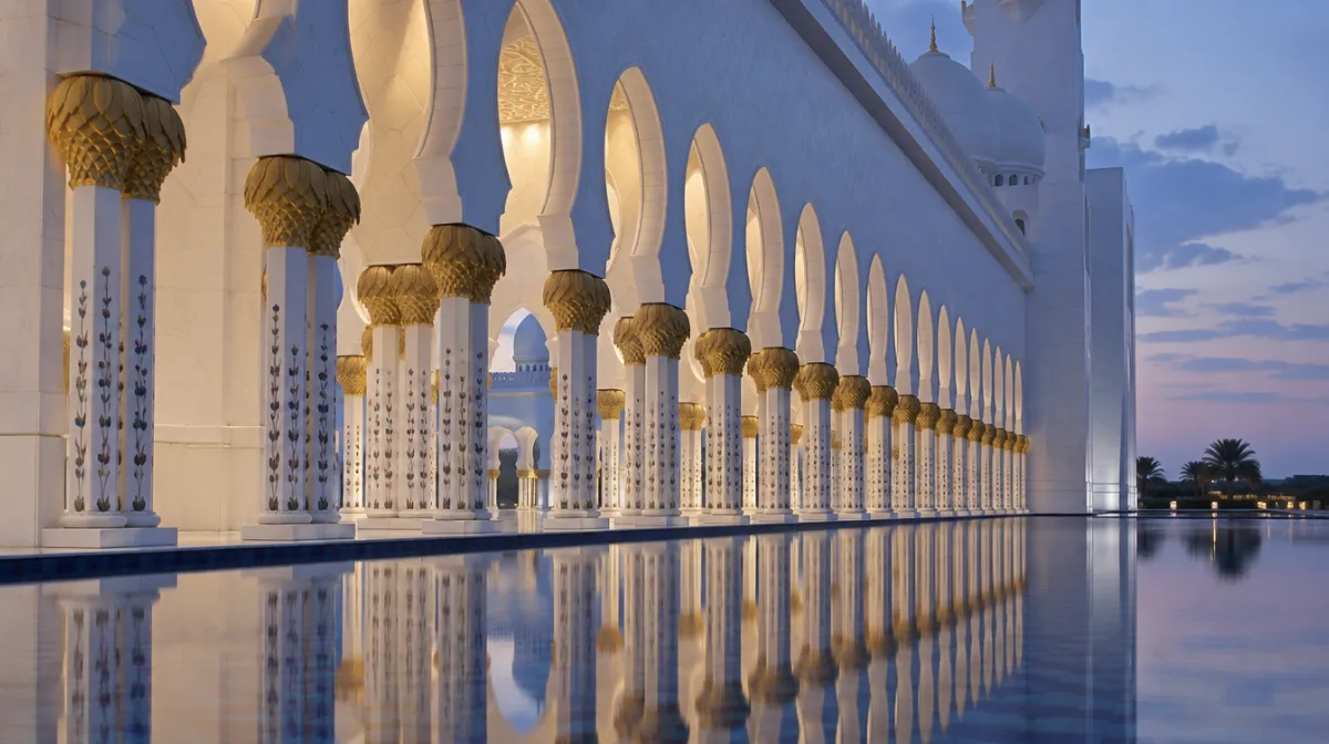 A white marble arcade with gold-topped columns reflected in a still pool at dusk at Sheikh Zayed Grand Mosque